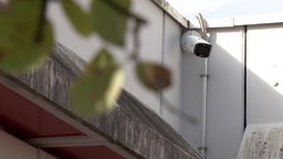 Auf dem Bild ist die Außenfassade einer Schule zu sehen. In einer Ecke unter dem Dach hängt eine Überwachungskamera.