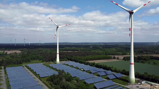 Zwei Windräder stehen auf einer freien Fläche, darunter Solaranlagen.