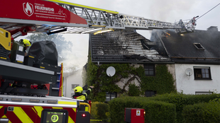 Feuerwehrleute auf einer Drehleiter löschen einen Brand, der aus dem Dachstuhl eines efeubewachsenen Hauses kommt.