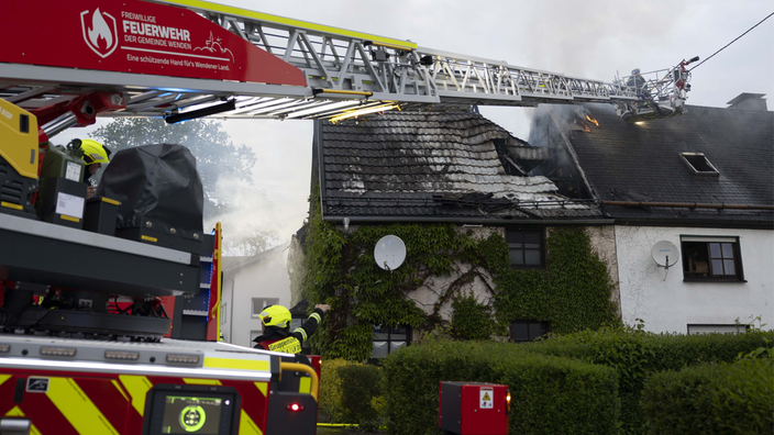 Feuerwehrleute auf einer Drehleiter löschen einen Brand, der aus dem Dachstuhl eines efeubewachsenen Hauses kommt.