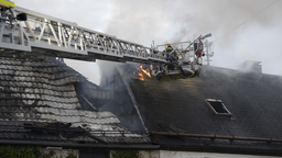 Flammen und Rauch kommen aus dem Dachstuhl eines Wohnhauses. Feuerwehrleute auf einer Drehleiter löschen den Brand. 