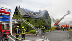 Feuerwehrleute in neongelben Helmen stehen vor einem brennenden Wohnhaus. Links und rechts stehen rote Feuerwehrautos mit Drehleitern.