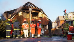 Feuerwehrleute stehen vor einem ausgebrannten Wohnhaus. 