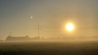 Sonnenaufgang im Nebel über einem Feld mit Strommasten im Hintergrund