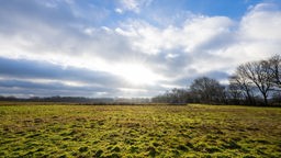 27.12.2022, Nordrhein-Westfalen, Warendorf: Vor einer grünen Wiese, scheint die Sonne durch weiß-graue Wolken und blauem Himmel hindurch