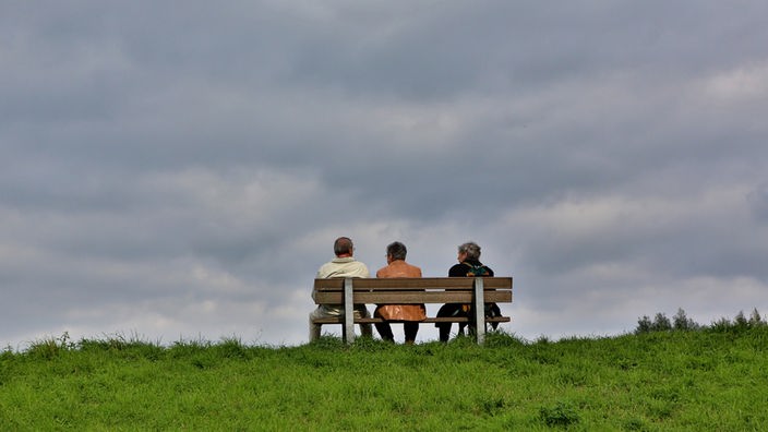 Drei Personen sitzen auf einer Bank, Himmel bewölkt