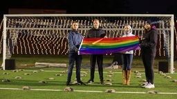 Vier Personen halten als Zeichen der Solidarisierung eine Regenbogenflagge vor einem Fußballtor hoch.