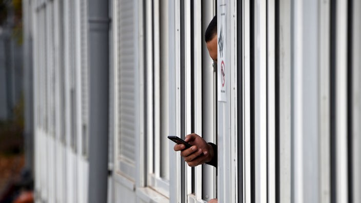 Ein Mann hält in einem Wohncontainer sein Smartphone aus dem Fenster. 