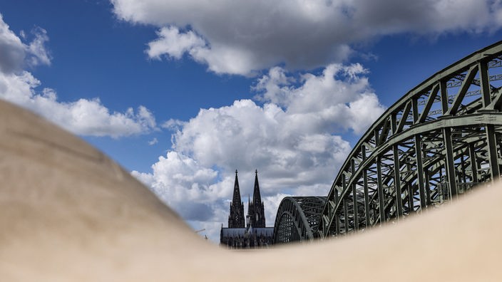 Wolken ziehen über Dom und Hohenzollernbrücke hinweg, die hinter rundlich geformten Sitzelementen zu sehen sind.