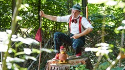 André Gatzke steht mit Dr. Kläver Vogelkuscheltier im Wald, umgeben von Bäumen.