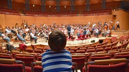 Ein kleiner Junge sitzt in den Reihen der Kölner Philharmonie und blickt auf das übende Sinfonieorchester.