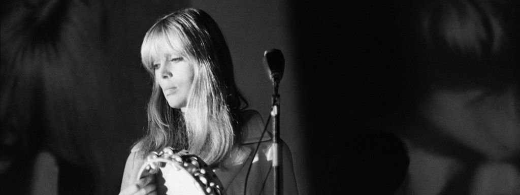 Christa Päffgen, bekannt als Sängerin Nico, steht mit einem Tamburin in der Hand vor einem Mikro und schaut traurig.