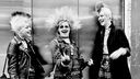 Drei weibliche Punks lachen gemeinsam, alle tragen Mohawk, die in der Mitte streckt der Kamera den Mittelfinger entgegen (London 1970er).