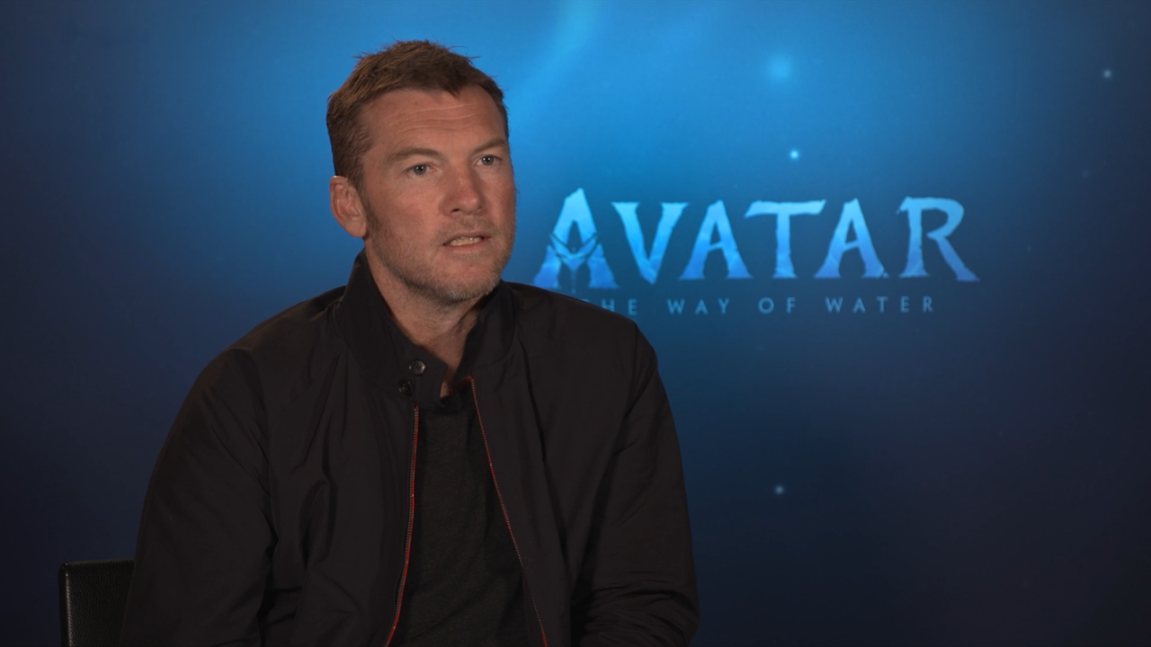 Schauspieler Sam Worthington sitzt vor einem blauen Hintergrund mit Avatar Schriftzug