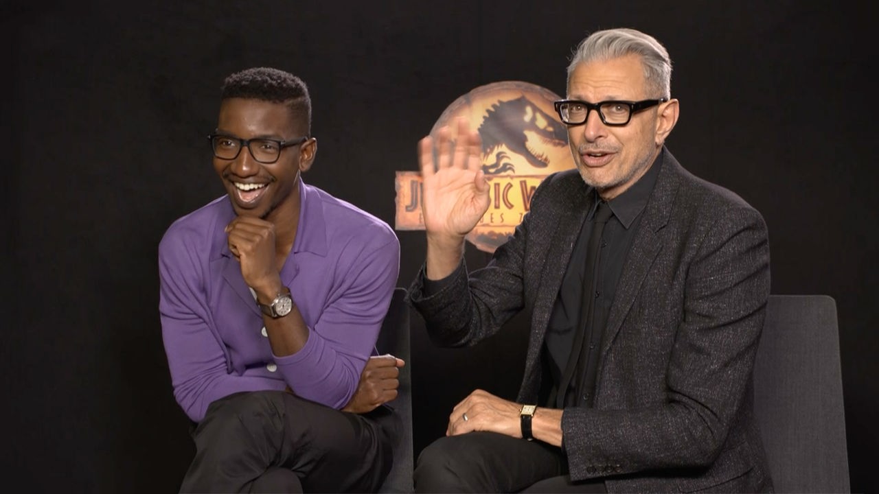 Jeff Goldblum und Mamoudou Athie sitzen vor einem schwarzen Vorhang. Im Hintergrund ist das Logo von Jurassic World zu sehen