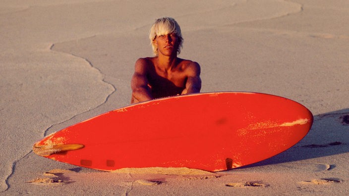 Der amerikanische Surfer Bunker Spreckels sitzt auf einem Strand vor seinem roten Surfbrett.