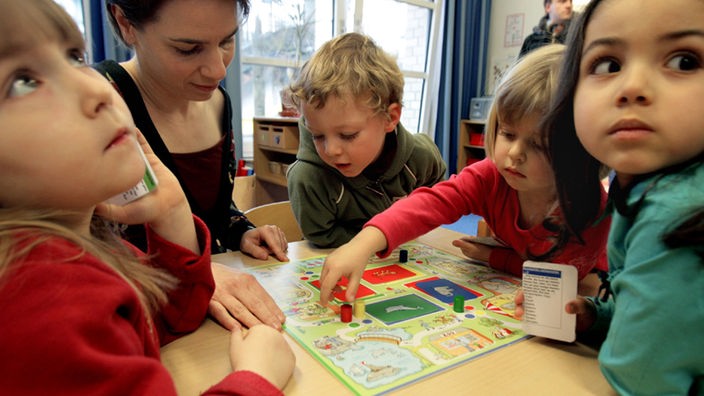 4 Kinder sitzen mit der Erzieherin im Kindergarten an einem Tisch und spielen ein Brettspiel