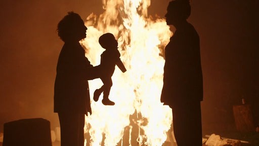 The Baby - Das Baby, gehalten von einer Frau zu einer anderen Frau hin vor einem lodernden Feuer.