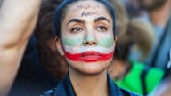 Junge, protestierende Frau mit den Nationalfarben des Irans geschminkt und mit dem Namen Amini auf der Stirn. 