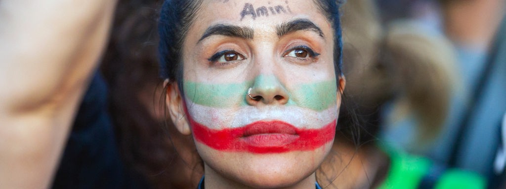 Junge, protestierende Frau mit den Nationalfarben des Irans geschminkt und mit dem Namen Amini auf der Stirn. 