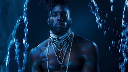 Albumcover - Jerome Thomas: "Submerge" - der Sänger mit nacktem Oberkörper und Perlenketten behangen steht zwischen Ketten. Die Atmosphäre dunkelblau, wie eine Unterwasserlandschaft. 
