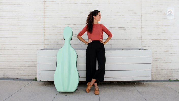 Die Musikerin Sarah Amiel steht neben ihrem Cello und schaut zur Seite.