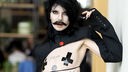 Der nicht binäre irische Sänger Bambie Thug steht lässig, mit abgeklebten Brustwaren, langen schwarzen Haaren und Schnäuzer für ein Foto modell. 