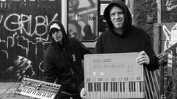 Inklusionsprojekt "Ick mach Welle" - schwarzweiß Foto: zwei Männer mit Synthesizer - davon einer gezeichnet, der andere real - zeigen die Instrumente in die Kamera. 