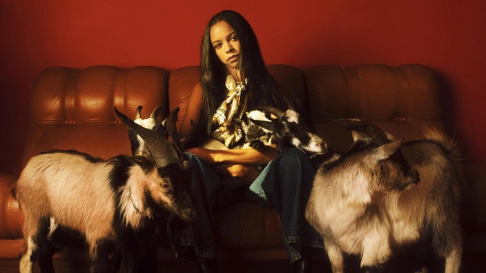 Cover des Albums "Capricorn Sun" von TSHA: Junge Frau sitzt auf einem Sofa; links und rechts von ihr steht je ein Bock