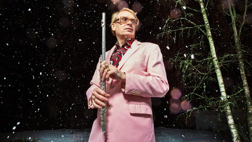 Der Musiker Jimi Tenor mit Querflöte, rosa Anzug und Goldrand-Brille im nächtlichen Schnee