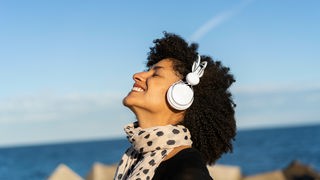 Eine Frau hört sich fröhlich Musik über Kopfhörer am Meer an.