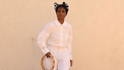 Santigold: "Shake" - die Sängerin, ganz in weiß gekleidet mit einem Tambourin in der Hand vor einer Wand stehend. 