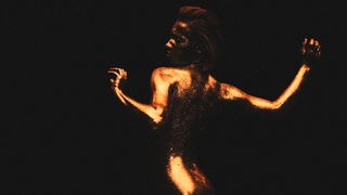 Videostill aus "Clit is good": Suzane nackt, mit goldfarbenem und glitzerndem Körper im fahlen Licht