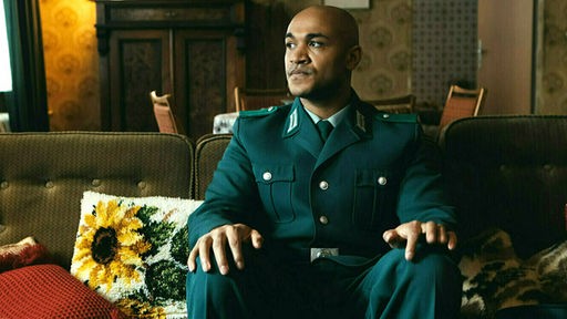Malick Bauer als Sam sitzt in Polizeiuniform auf einem Sofa. Szene des Films "Sam – Ein Sachse"