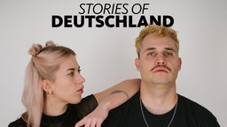 Eine Mann und eine Frau stehen vor einer weißen Wand, die Frau lehnt sich aufrecht an die Schulter des Mannes, dazu das Logo von Stories of Deutschland.