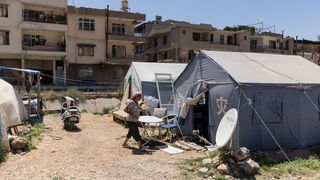 Eine Frau geht zwischen ihrem Zelt und einem Beet in einer Zeltstadt im Zentrum von Antakya, wo sie im Freien versucht, die Hitze zu ertragen