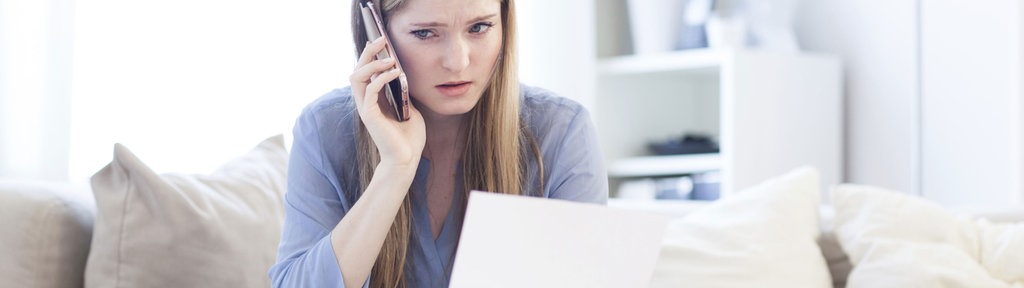 Eine Frau am Handy, sitzend auf einem Sofa. In der Hand hält sie Unterlagen