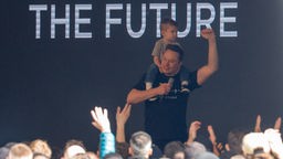 Elon Musk con il figlio sulle spalle mentre viene applaudito dai dipendenti della fabbrica