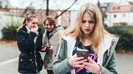 Devojka u školskom dvorištu uplašeno gleda u Smartphone, iza nje se dve devojke smeju