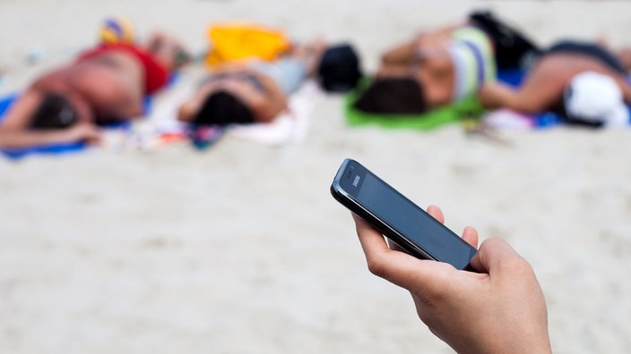 Ruka na plaži drži mobilni telefon, u pozadini se vide ljudi koji se sunčaju