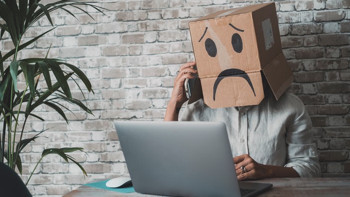 Čovek na kompjuteru sa telefonskom slušalicom, na glavi mu kartonska kutija sa nacrtanim tužnim izrazom lica