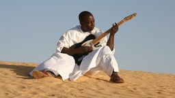 Vieux Farka Touré mit einer E-Gitarre auf einer Düne sitzend