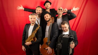 Die Band Les Yeux d'la Tête steht mit Instrumenten vor einem roten Hintergrund.