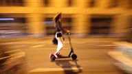 Eine junge Frau fährt abends auf einem E-Scooter durch die Stadt, der Hintergrund ist verschwommen.