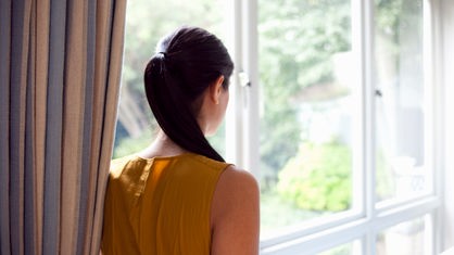Eine Frau mit dunklem Pfereschwanz steht vor einer großen Fensterfront und schaut hinaus.