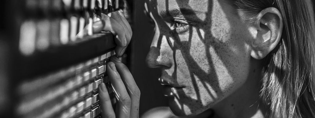 Schwarz-weiß: Eine Frau schaut durch einen engmaschigen Zaun, auf ihr Gesicht fallen die Schatten der Maschen.
