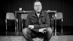 Der Dramatiker iund Regisseur René Pollesch sitzt auf einer Theaterbühne, das Bild ist in schwarz-weiß.