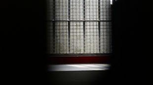 Blick durch ein freies Schlüsselloch in eine Zelle mit vergittertem Fenster.