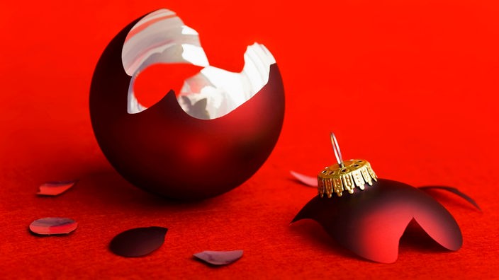 Eine rote Weihnachtskugel liegt zebrochen auf dem Boden, vor rotem Hintergrund.