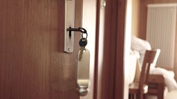 In einer offenen Tür eines Hotelzimmers steckt ein Schlüssel, vom Zimmer ist ein Stuhl und Bettwäsche zu erkennen.